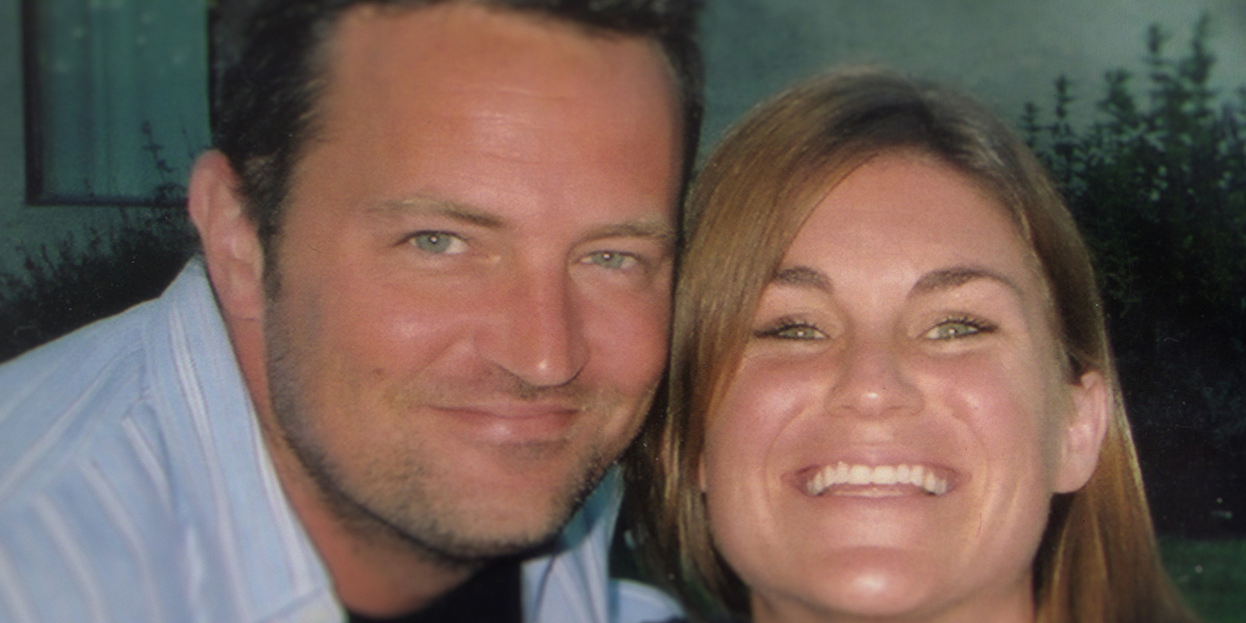 "Two people smiling closely together outdoors in soft light, with greenery and a window in the background."